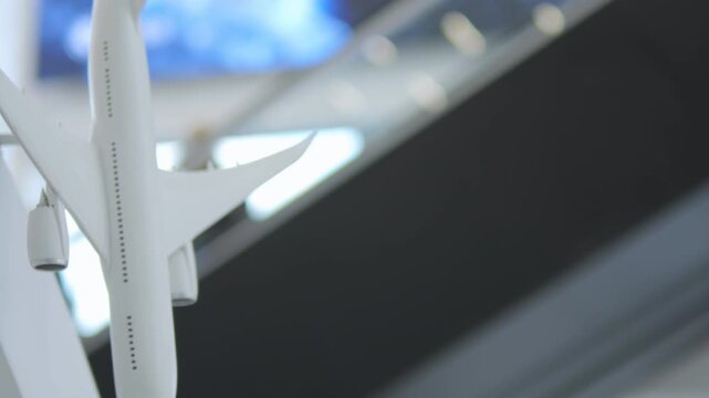 White airplane model displayed indoors with soft lighting. Background shows blurred ceiling and structural beams. Camera slowly moves right, focusing on aircraft's wing and fuselage