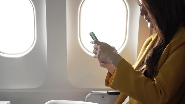 Young woman in airplane window seat using smartphone, natural light, travel and connectivity concept, close-up side view with copy space.