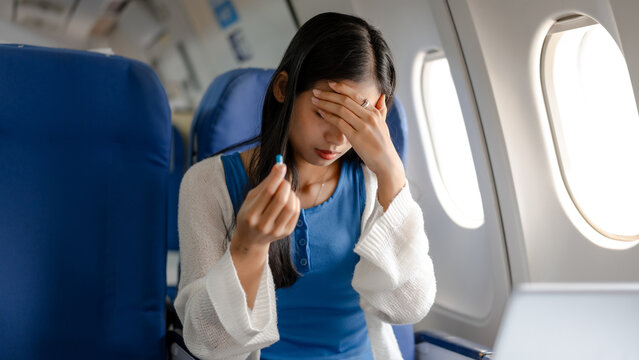 Young woman passenger in airplane seat holding pill and covering forehead, looking sick with headache or nausea, travel illness concept, window light, copy space.