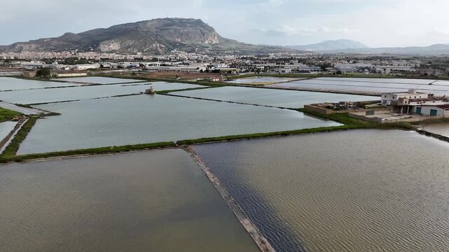 Drone panorama of Salina Galia Canino salt pans near Trapani, Sicily, with mountain skyline, cloudy sky, city edge, and coastal basins.