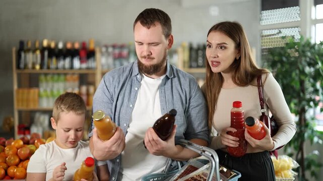 Attentive couple and their little boy deciding what juice to buy during shopping in supermarket 