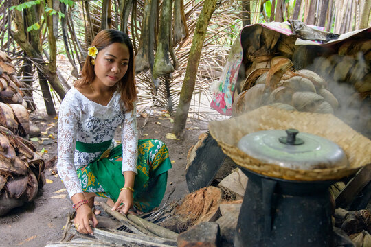 Balinese woman in white lace kebaya and green batik tending traditional wood fire stove with steaming pot surrounded by coconut shells