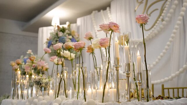 Roses and candles are arranged on a table at an event location. The background features decorations with fabric and lights. The setup is ready for a celebration.