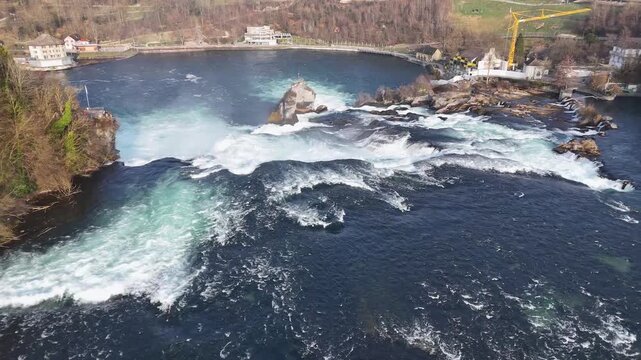 Backlit aerial view of Rhine Falls with mist, rushing water, bridge arches, and riverside buildings in Switzerland.
