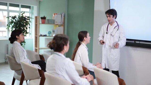 Young male doctor in medical gown with remote control gives presentation to colleagues doctors in conference room