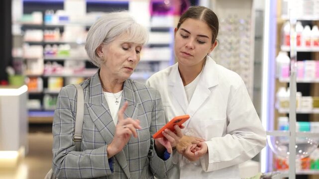 Pharmacist girl communicates with mature female client, looks at mobile phone screen, helps to identify desired drug in photo