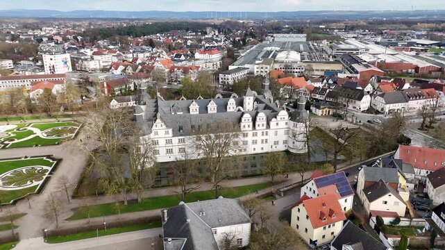 Wide aerial panorama of Schloss Neuhaus, landscaped garden, moat, parkland, and cloudy sky in Paderborn, North Rhine-Westphalia, Germany.