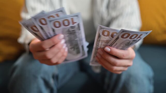 A person sits on a couch and counts a stack of cash with bills visible in their hands. The activity suggests financial management and the handling of money indoors