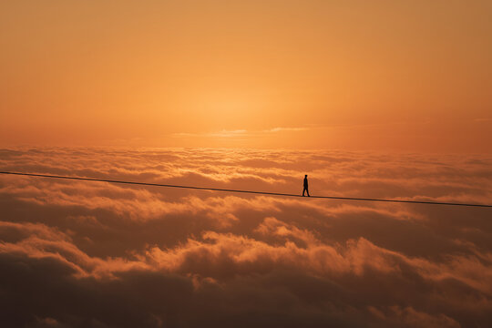 An aerial view of a person walking on a tightrope or scrambling above a vast sea of ​​clouds amidst a sunset.