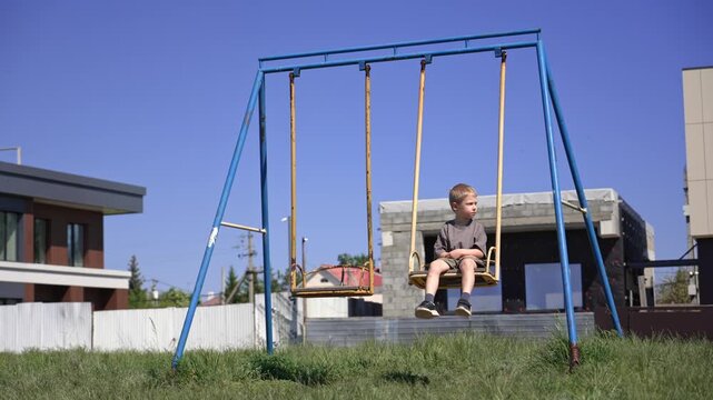 Unhappy lonely kid sitting on a rusty swing in a neglected backyard playground, feeling bored and sad during a sunny summer day, with modern houses and construction in the background