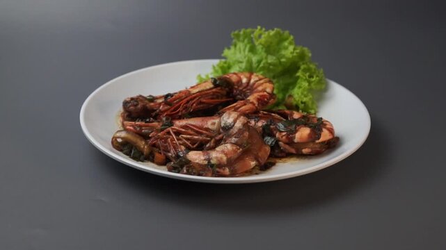 Hand holding a white plate of stir-fried giant tiger prawns in sweet soy sauce with lettuce garnish. Top view commercial shot of serving a seafood dish isolated on grey background.