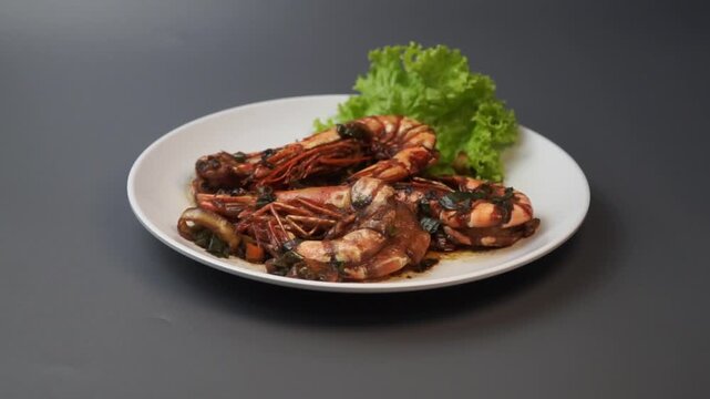 Hand holding a white plate of stir-fried giant tiger prawns in sweet soy sauce with lettuce garnish. Top view commercial shot of serving a seafood dish isolated on grey background.