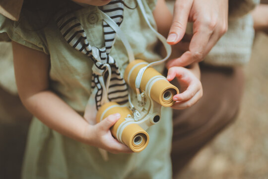 Close-up of a young Asian toddler girl holding yellow toy binoculars around her neck, learning to use them with the gentle guidance of a parent&rsquo;s hands during an outdoor family adventure and education