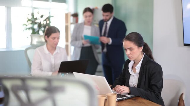  Young woman sits at table and works on laptop in group of colleagues in office