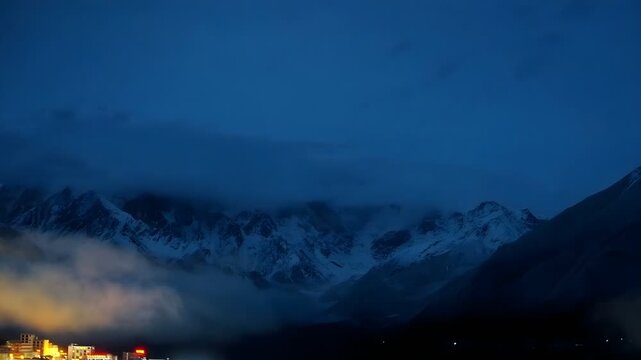 Stunning night time lapse of the sacred Namcha Barwa snow peak with moving clouds and an illuminated Tibetan village at the base in Tibet China.