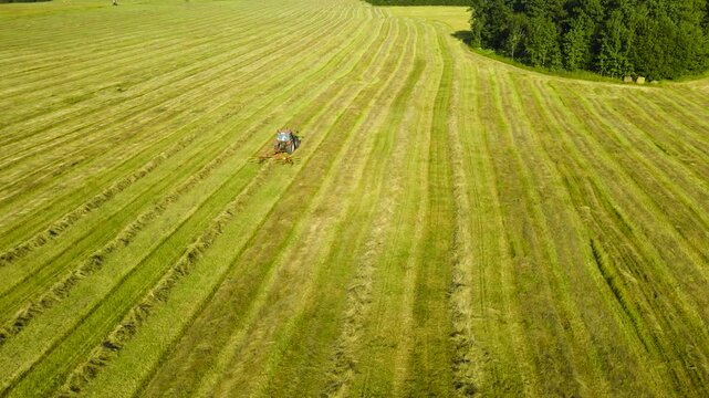 Agricultural tractor with a hay tedder working in a freshly mown field. Aerial perspective of seasonal farm work preparing animal fodder
