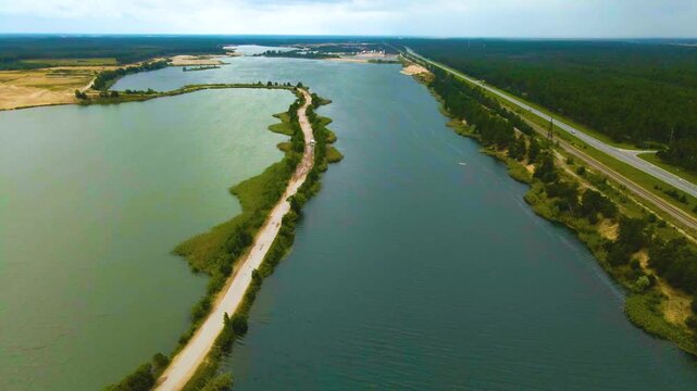 Aerial drone footage flying over a sand quarry transformed into artificial lakes. A narrow, winding road separates the bodies of water
