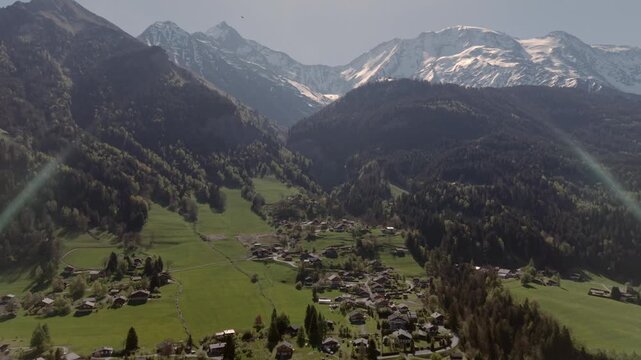 FPV aerial view of a mountain village nestled in a lush valley with snow-capped peaks and dense forests in Saint-Gervais-les-Bains, Auvergne-Rhone-Alpes, France.