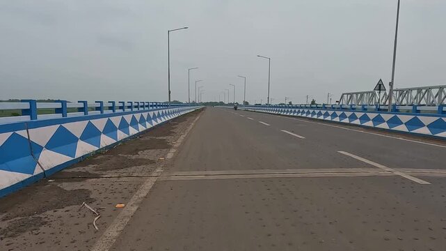 Wide-angle view of a modern concrete highway bridge leading towards Kirnahar, featuring distinct blue and white geometric painted side railings under an overcast cloudy sky with minimal motorcycle tra