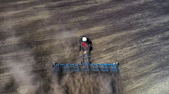 Watch as a vibrant red tractor gracefully plows through a golden field. The dust clouds billow in its wake, showcasing the raw beauty of agricultural life from above. Aerial FHD footage.