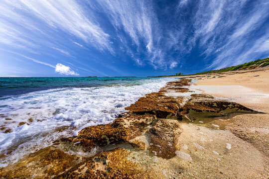 Dramatic tropical shoreline at Green Turtle Cay, Bahamas, with turquoise Caribbean waves washing over rocky limestone formations under a vibrant blue sky filled with wispy clouds.
