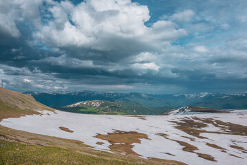 Sunlit snowfields and thawed ground on hill top with view to green hills and big snowy mountain range far away under cloudy sky. Dramatic landscape with sunlight and shadows of clouds high mountains. © Daniil