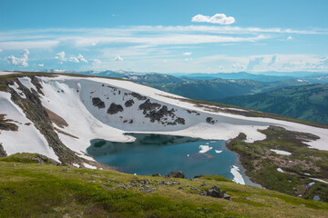 Scenic sunlit landscape with alpine lake in rocky snowy cirque near stone hill top during thaw. Ice floats in mountain lake among rocks and snows with view to forest mountain range under cloudy sky. © Daniil
