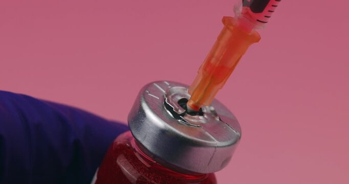 Syringe needle penetrating the rubber stopper of a medical vial held by a hand in a detailed view on a pink gradient background.