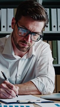 Auditor scrutinizing financial documents at desk, careful pen annotations, glasses reflecting charts, focused expression, shelves of binders behind, methodical verification and number checking