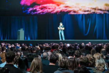 Naklejka premium Keynote speaker addresses large audience during conference presentation on stage, colorful screen above, crowded theater atmosphere.