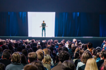 Naklejka premium Large audience watching speaker on stage during conference presentation, focused on crowd engagement and public speaking.