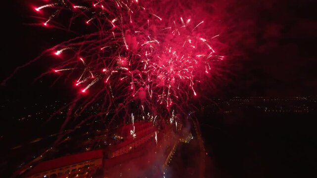 FPV aerial view of red fireworks exploding over the city lights and buildings at night in Novi Sad, Vojvodina, Serbia.