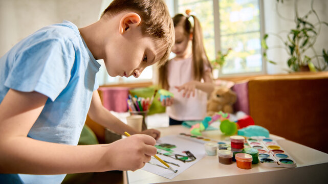 Focused boy painting while girl plays with modeling clay in bright creative room. Concept of kids creativity, art practice, independent play, education content, and craft supplies.