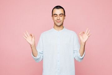 Young spiritual middle eastern man wear casual blue cotton shirt hold hands in yoga om aum gesture relax meditate try calm down isolated on plain pastel light pink wall background. Lifestyle concept. © ViDi Studio