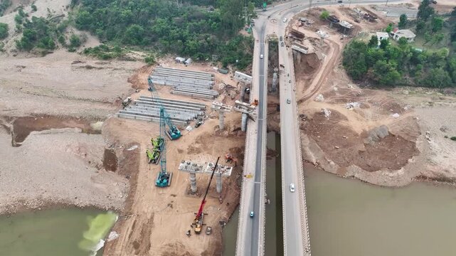 Heavy cranes lifting concrete girders at bridge construction site. High-angle aerial drone footage capturing the construction of a massive new highway bridge.