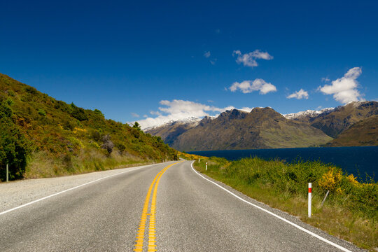 A view along a road with yellow double lines leading to snow capped mountains beside a blue lake.