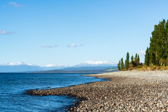 Lake shore with pebble beach, trees and snow capped mountains in the background