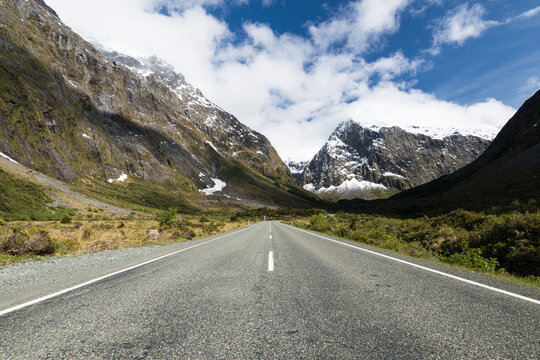 A view of a road with a central dotted white line, leading to snow capped mountains