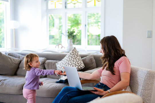 Toddler girl giving toy aeroplane to mum sitting on the couch holding a laptop in the living room