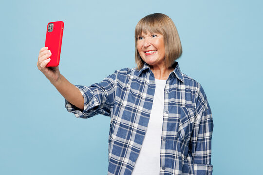 Elderly blonde woman 50s years old wear checkered shirt casual clothes doing selfie shot on mobile cell phone post photo on social network isolated on plain pastel blue background. Lifestyle concept.