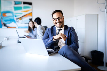 Obraz premium Confident businessman in suit smiling at camera, working on laptop in modern office, colleagues blurring in background
