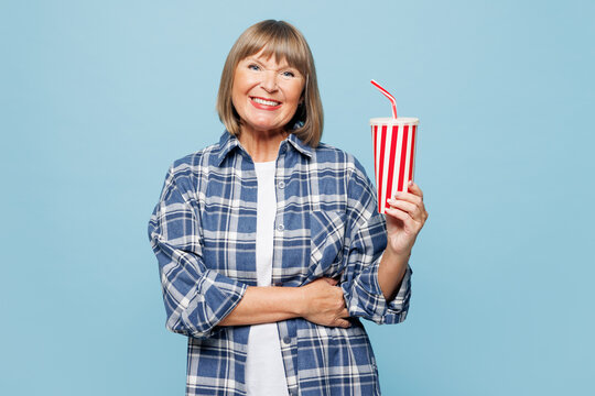 Elderly smiling happy fun blonde woman she wear checkered shirt casual clothes hold in hand cup of soda pop cola fizzy water isolated on plain pastel blue background studio portrait Lifestyle concept