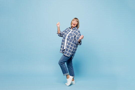 Full body elderly blonde woman 50s years old wear checkered shirt casual clothes doing winner gesture celebrate clench fists say yes raise up leg isolated on plain blue background. Lifestyle concept.