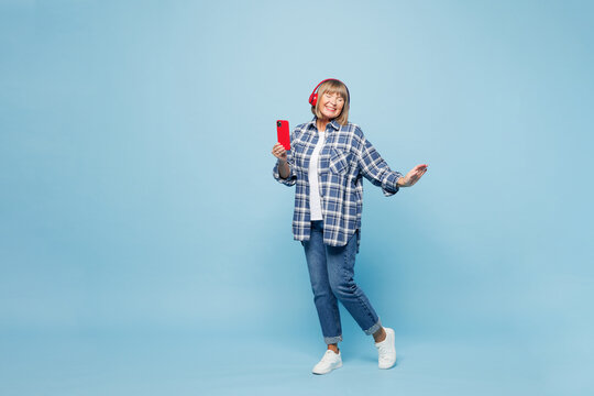 Full body elderly blonde woman wear checkered shirt casual clothes listen to music in headphones hold use mobile cell phone isolated on plain pastel blue background studio portrait. Lifestyle concept.