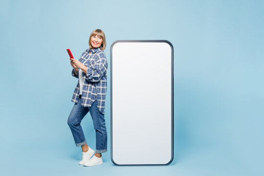 Full body smiling elderly blonde woman 50s years old wear checkered shirt casual clothes stand near big huge blank screen mobile cell phone with area use smartphone isolated on plain blue background.