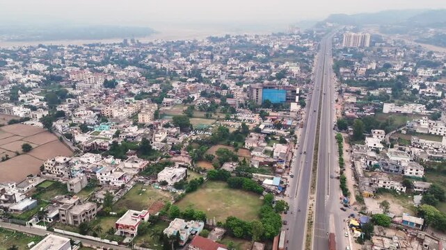 Aerial view of major highway corridor through dense Indian city outskirts. The wide establishing shot illustrates the intersection of heavy regional transportation and daily suburban life.