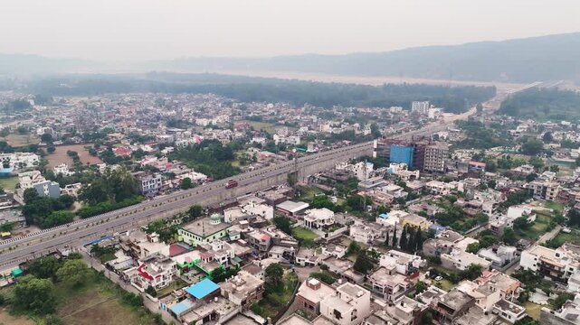 Aerial view of major highway corridor through dense Indian city outskirts. The wide establishing shot illustrates the intersection of heavy regional transportation and daily suburban life.