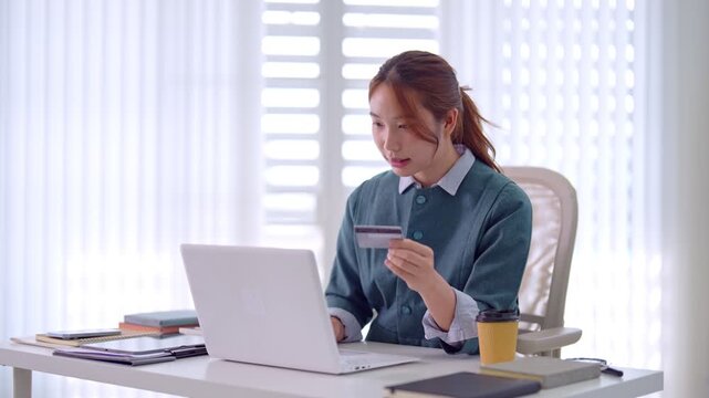 young Asian businesswoman using credit card laptop computer online shopping payment ecommerce transaction home office desk modern workspace bright natural