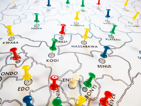 A high-angle shot of a white Nigerian map dotted with multi-colored push pins on state targets like Kogi and Abuja. The colorful pins create a vibrant, busy concept for logistics and demographics.