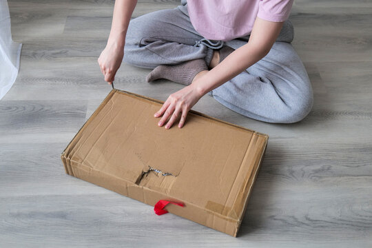 A woman is sitting on the floor at home and carefully opening a cardboard box with a knife, representing online shopping delivery or unpacking